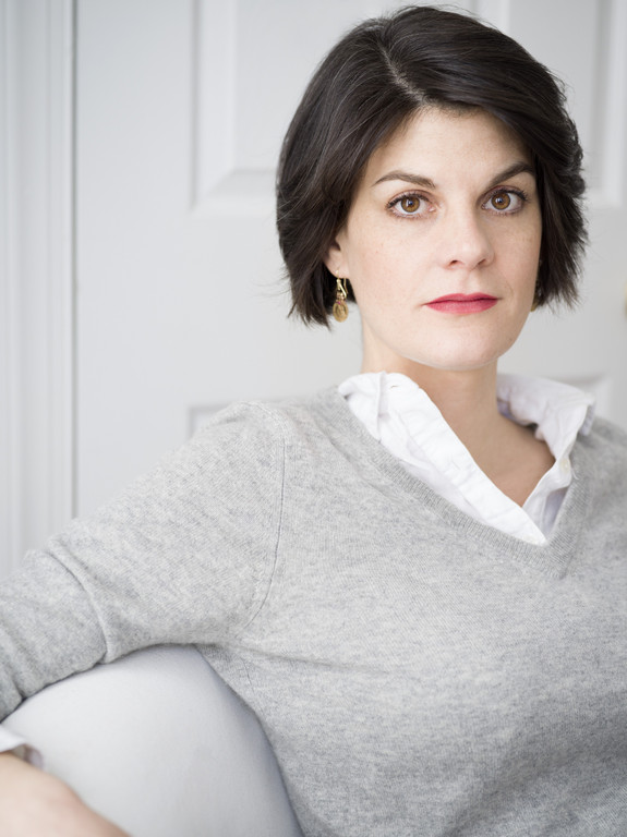 Das Bild zeigt die Autorin Nora Krug, eine Frau Mitte 40, die auf einem Sofa sitzt und offen und direkt in die Kamera blickt. Sie hat halblanges braunes Haar und trägt eine weiße Bluse unter einem grauen Pullover.