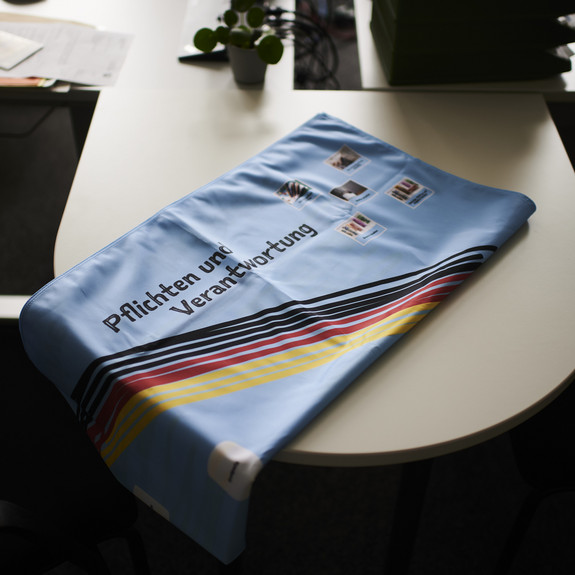 Auf einem Tisch liegt ein hellblaues Tuch mit der Aufschrift "Pflichten und Verantwortung". Unter dem Schriftzug sind schwarze, rote und gelbe Linien.
