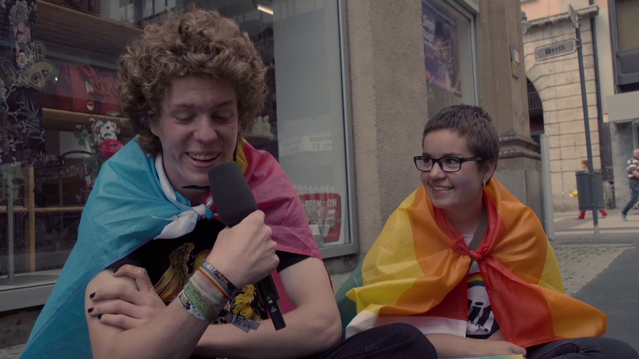 Man sieht zwei Personen mit Regenbogenfahnen, die interviewt werden