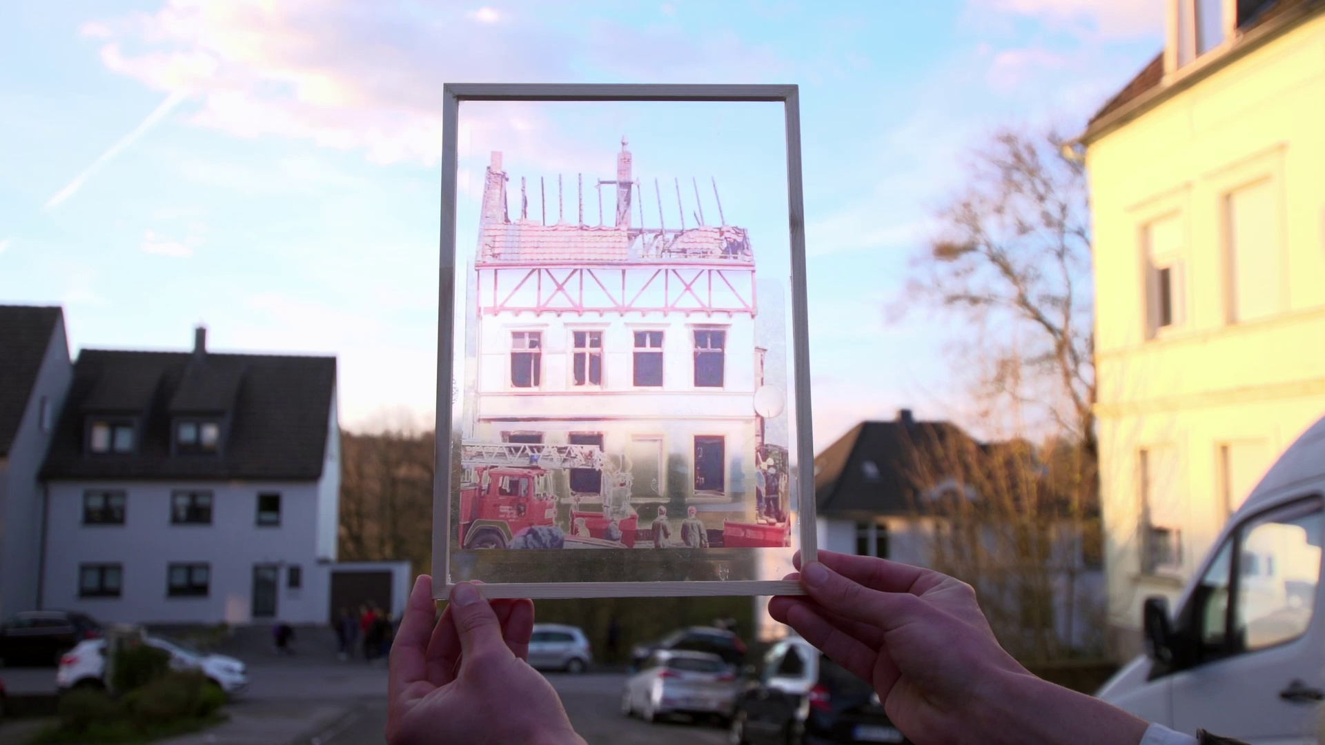 Filmscene: Zwei Hände halten ein Foto des ausgebrannten Hauses der Familie Genç in die Lücke auf der Unteren Wernerstraße 81 in Solingen, wo das Haus vor dem Brandanschlag gestanden hat.