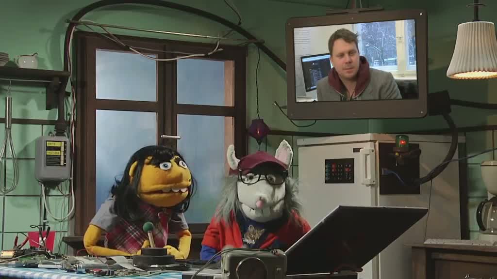 Zwei Handpuppen am Computer, im Hintergrund Moderator über TV-Screen.