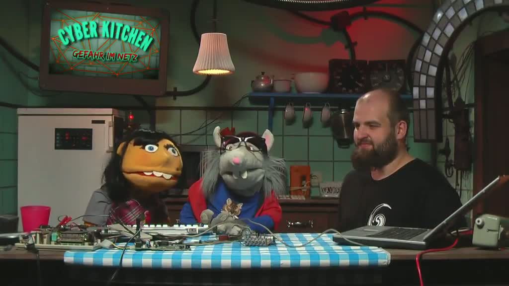 Zwei Handpuppen und Mann am Tisch mit technischen Geräten.