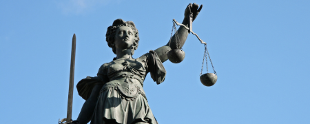 Das Bild zeigt die Figur der Justizia als Steinstatue von blauem Himmel. Sie hat mit einem Tuch verbundene Augen und hält in der linken Hand die ausbalancierte Waage des Rechts, in der rechten Hand ein Schwert.